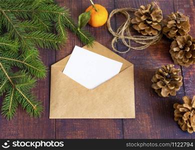 Blank white paper card in a brown envelope from handmade craft paper on an old wooden table in a Christmas style. Fir branches, mandarin and cones are on the table. Christmas and New Year.. Blank white paper card in a brown handmade envelope on an old wooden table in a Christmas style. Fir branches, mandarin and cones are on the table. Christmas and New Year.