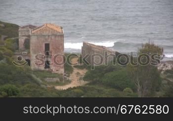 Blick auf ein altes Haus direkt am Ufer von einem Meer mit Wellen.