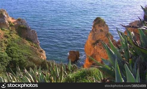 Blick auf Felsengebilde / Stein im blauen Meer von einem grnn bewachsenen Felsen; die Sonne spiegelt sich im Wasser. Knste der Algarve, Portugal.
