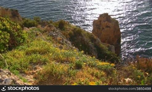 Blick auf Felsengebilde / Stein im blauen Meer von einem mit bunten Blumen / Gras bewachsenen Felsen; die Sonne glitzert / spiegelt sich im Wasser. Knste der Algarve, Portugal.