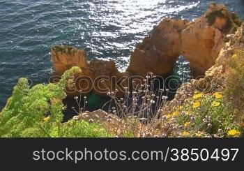 Blick auf Felsengebilde / Steine im blauen Meer von einem mit bunten Blumen bewachsenen Felsen; die Sonne glitzert / spiegelt sich im Wasser. Knste der Algarve, Portugal.