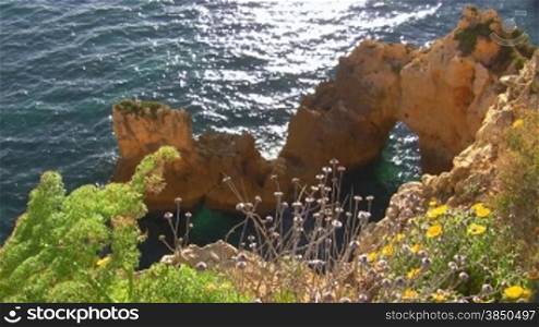 Blick auf Felsengebilde / Steine im blauen Meer von einem mit bunten Blumen bewachsenen Felsen; die Sonne glitzert / spiegelt sich im Wasser. Knste der Algarve, Portugal.