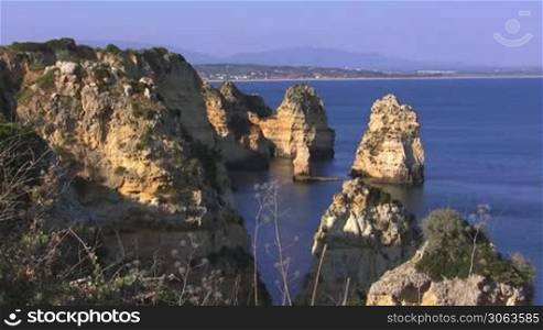 Blick auf Felsengebilde / Steine / Steininseln, teilweise grun bewachsen, die aus dem blauen Meer ragen; im Hintergrund die Kuste der Algarve, Portugal und Berge.
