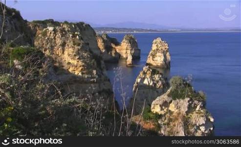 Blick auf Felsengebilde / Steine / Steininseln, teilweise grun bewachsen, die aus dem blauen Meer ragen; im Hintergrund die Kuste der Algarve, Portugal und Berge.