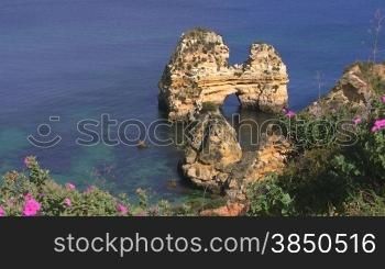 Blick auf Felsengebilde / Steininseln im blauen Meer von einem grnn bewachsenen Felsen mit rosa Blumen; Knste der Algarve, Portugal.