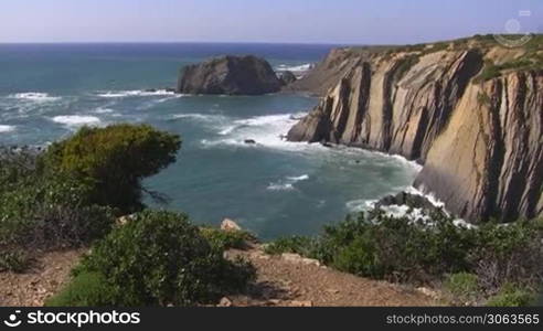 Blick auf hohe Klippen / das Meer von einem Aussichtspunkt mit grunen Buschen; Felsen im Meer, Kuste der Algarve in Portugal; blauer Himmel.