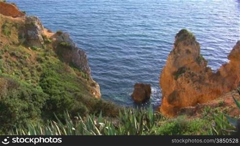 Blick von mit grnnen Bnschen, Gras und Kakteen bewachsenen Felsen auf das Meer und Felsengebilde / Steine / Steininseln, die aus dem blauen Meer ragen; im Hintergrund die Knste der Algarve, Portugal und Berge.