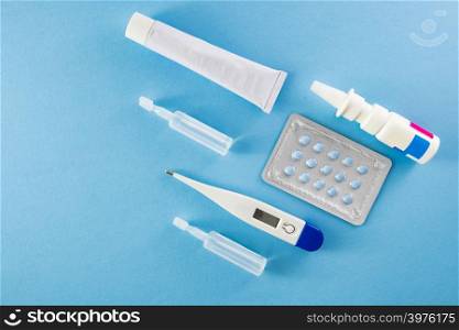 Blister pack of blue pills, ampoules, nasal spary, white ointment tube and digital thermometer on blue background- healthcare and medical concept
