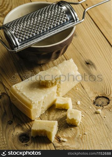 Block of parmesan cheese on a wooden table with grater in the background.