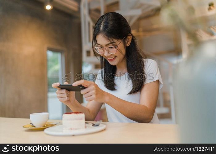 Blogger Asian friendly women influencer eat cake at night cafe. Beautiful young lady happy relax fun using technology mobile phone taking a photo of her food upload in social media at college campus.