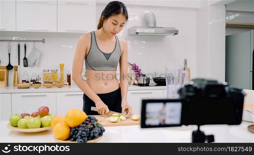Blogger sporty Asian woman using camera recording how to make apple juice video for her subscriber, female use organic fruit making apple juice by herself at home. Healthy food concept.