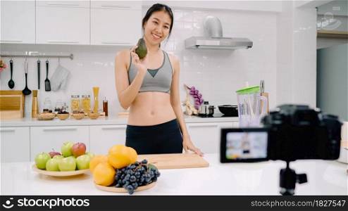 Blogger sporty Asian woman using camera recording how to make avocado juice video for her subscriber, female use organic fruit making avocado juice by herself at home. Healthy food concept.