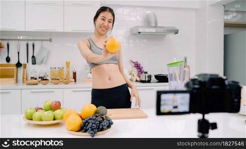 Blogger sporty Asian woman using camera recording how to make orange juice video for her subscriber, female use organic fruit making orange juice by herself at home. Healthy food concept.