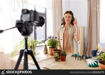 blogging, gardening and housework concept - happy woman or blogger with camera and pot flowers recording tutorial video and pointing finger up at home. happy woman or blogger planting flowers at home