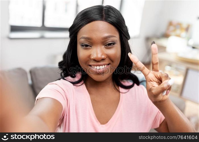 blogging, gesture and people concept - happy smiling african american woman taking selfie and peace hand sign at home. happy african american woman taking selfie at home