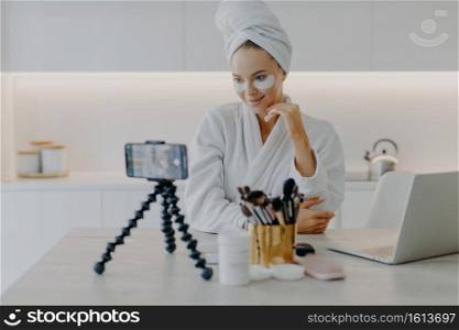 Blogging modern technologies skin care and beauty concept. Pleased beautiful woman records video tutorial at home applies beauty pads under eyes shoots broadcast live video to social network