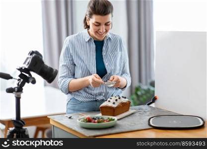 blogging, photographing and people concept - happy smiling female photographer or food blogger with camera pouring powdered sugar to cake in kitchen at home. food blogger with camera icing cake in kitchen