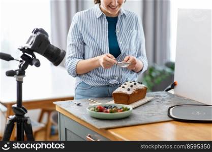 blogging, photographing and people concept - happy smiling female photographer or food blogger with camera pouring powdered sugar to cake in kitchen at home. food blogger with camera icing cake in kitchen