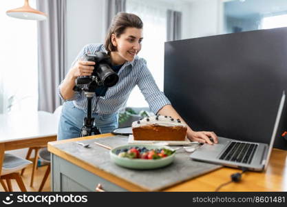 blogging, profession and people concept - happy smiling female food photographer with laptop computer and camera photographing cake in kitchen at home. food photographer with camera working in kitchen