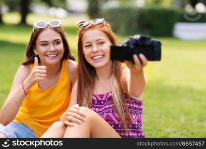 blogging, technology and leisure concept - teenage girls, friends or bloggers recording video by camera in summer park and showing thumbs up. teenage bloggers recording video by camera in park