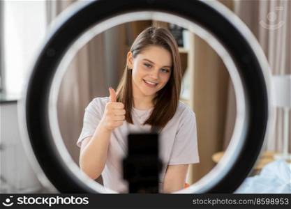 blogging, technology and people concept - happy smiling girl blogger with ring light and smartphone streaming at home and showing thumbs up gesture. girl blogger with ring light streaming at home