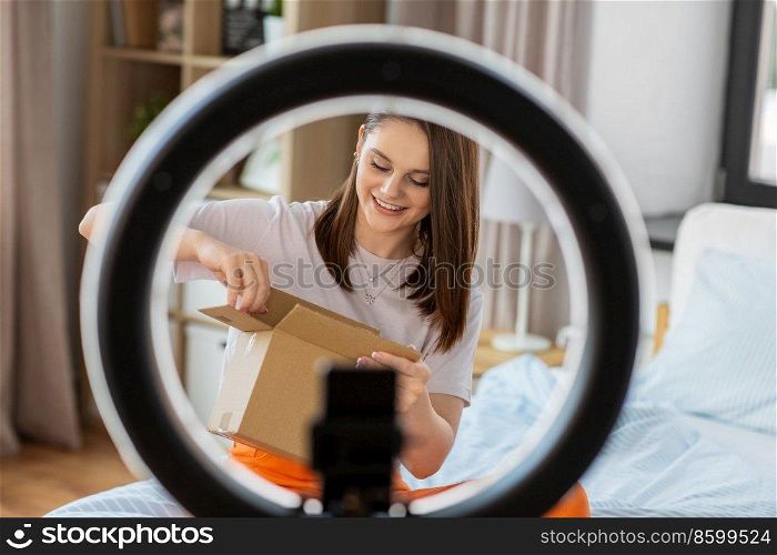 blogging, technology and people concept - happy smiling girl blogger with ring light and smartphone unpacking parcel box at home. happy girl blogger unpacking parcel box at home