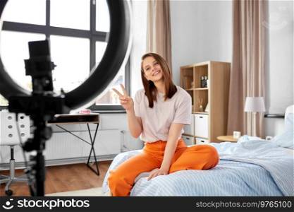 blogging, technology and people concept - happy smiling girl blogger with ring light and smartphone showing peace gesture at home. girl blogger with ring light streaming at home