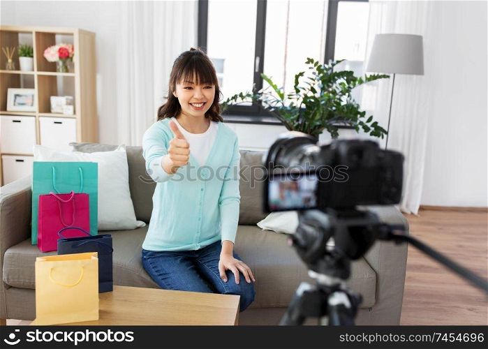 blogging, technology, videoblog and people concept - happy smiling asian woman or blogger with shopping bags recording video blog by camera and showing thumbs up gesture at home. female blogger making video blog about shopping