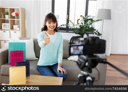 blogging, technology, videoblog and people concept - happy smiling asian woman or blogger with shopping bags recording video blog by camera and showing thumbs up gesture at home. female blogger making video blog about shopping