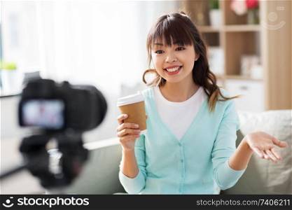 blogging, technology, videoblog and people concept - nice asian woman or blogger with camera and takeaway coffee cup recording video blog at home in morning. asian female blogger with coffee recording video