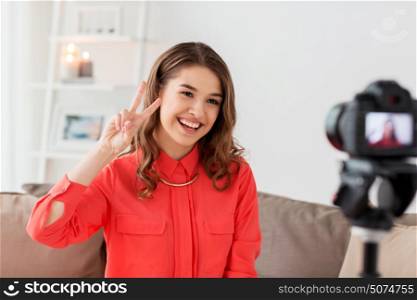 blogging, technology, videoblog, mass media and people concept - happy smiling woman or blogger with camera recording video and showing peace hand sign at home. woman with camera recording video at home