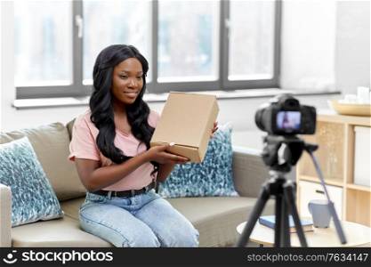 blogging, videoblog and people concept - african american female video blogger with parcel box and camera videoblogging at home. female video blogger with camera and box at home