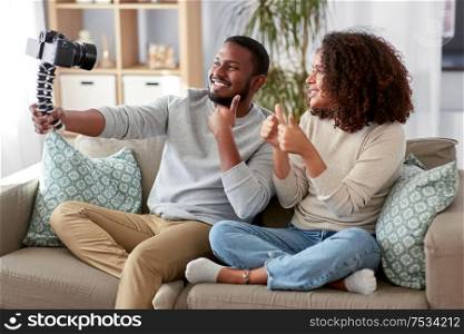blogging, videoblog and people concept - happy african american couple of video bloggers with camera videoblogging at home and showing thumbs up. couple of video bloggers with camera at home