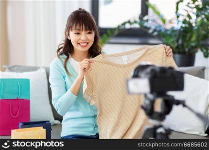 blogging, videoblog and shopping concept - happy smiling asian woman or fashion blogger with t-shirt, shopping bags and camera recording video blog at home. female fashion blogger making blog about shopping