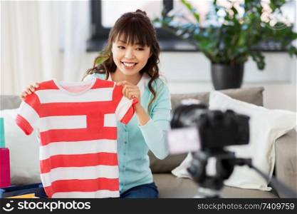 blogging, videoblog and shopping concept - happy smiling asian woman or fashion blogger with t-shirt, shopping bags and camera recording video blog at home. female fashion blogger making blog about shopping