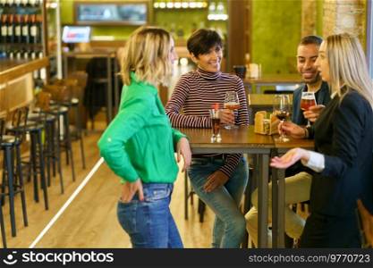 Blond female with glass of wine telling story to diverse male and female friends while gathering around table in bar together. Blond woman speaking with friends in pub
