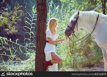 Blond girl feed grass to her white horse. Blond girl feed grass to her white horse in a magic light forest near river