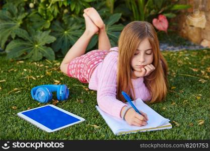 Blond kid girl homework lying on grass turf writting notebook