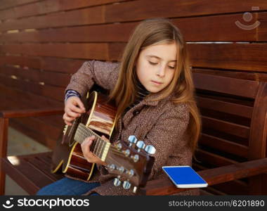 blond kid girl learning play guitar with smartphone winter beret on wooden background