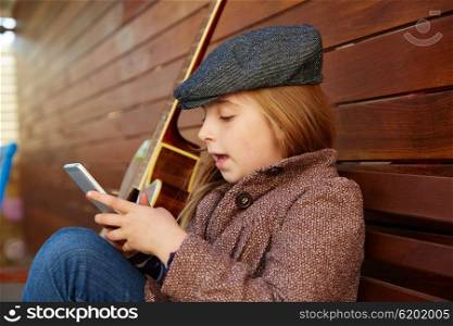 blond kid girl playing smartphone winter beret and guitar on wooden background