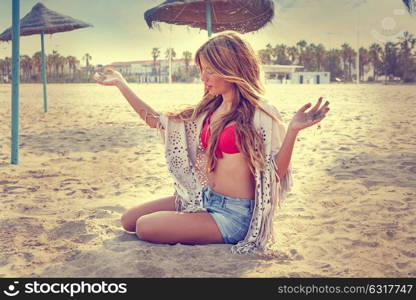 Blond teen girl sit on the beach sand near thatch umbrellas