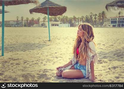 Blond teen girl sit on the beach sand near thatch umbrellas filtered image