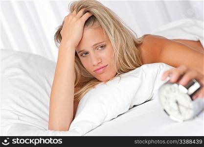 Blond woman holding alarm clock lying in white bed
