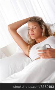 Blond woman holding white mug with coffee lying on white bed
