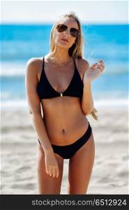 Blond woman in swimwear on the beach with aviator sunglasses. Young blonde woman with beautiful body in swimwear on a tropical beach with aviator sunglasses. Caucasian female with straight long hairstyle wearing black bikini.