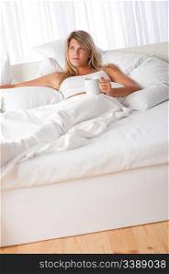 Blond woman lying in white bed drinking coffee relaxing