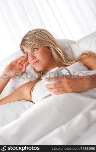 Blond woman lying in white bed holding cup of coffee
