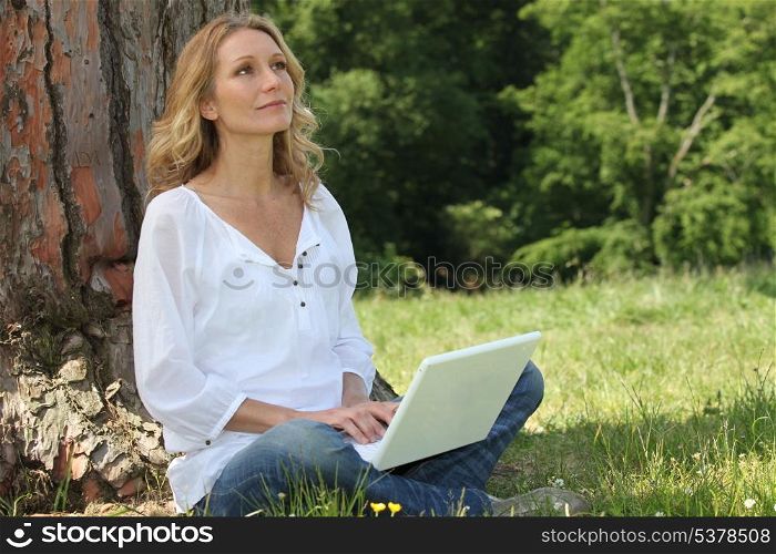 Blond woman sat by tree with laptop deep in thought