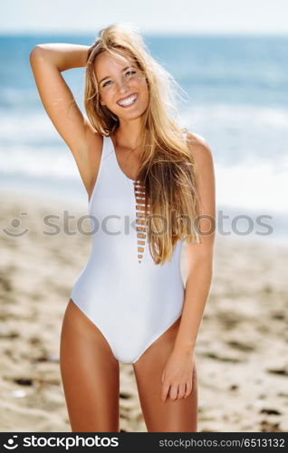 Blond woman with beautiful body in swimswit on a tropical beach. Young blonde woman with beautiful body in white swimsuit on a tropical beach. Caucasian female with straight long hairstyle smiling.