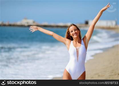 Blond woman with beautiful body in swimswit on a tropical beach. Young blonde woman with beautiful body in white swimsuit on a tropical beach with open arms. Caucasian female with straight long hairstyle smiling.